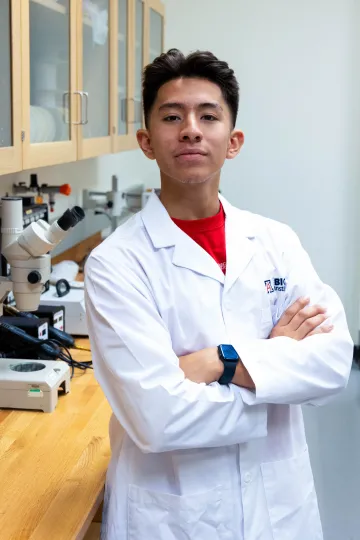 Student in a white lab coat stands confidently with crossed arms in a laboratory setting, smiling at the camera, with a microscope and other scientific equipment in the background.