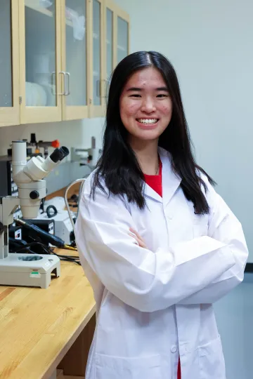 Student in a white lab coat stands confidently with crossed arms in a laboratory setting, smiling at the camera, with a microscope and other scientific equipment in the background.