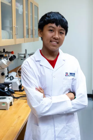 Student in a white lab coat stands confidently with crossed arms in a laboratory setting, smiling at the camera, with a microscope and other scientific equipment in the background.