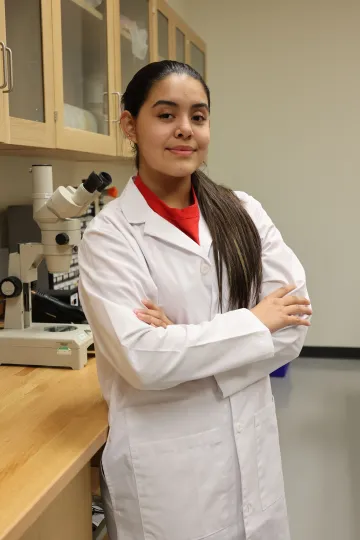 Student in a white lab coat stands confidently with crossed arms in a laboratory setting, smiling at the camera, with a microscope and other scientific equipment in the background.