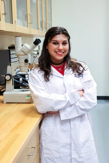 Student in a white lab coat stands confidently with crossed arms in a laboratory setting, smiling at the camera, with a microscope and other scientific equipment in the background.