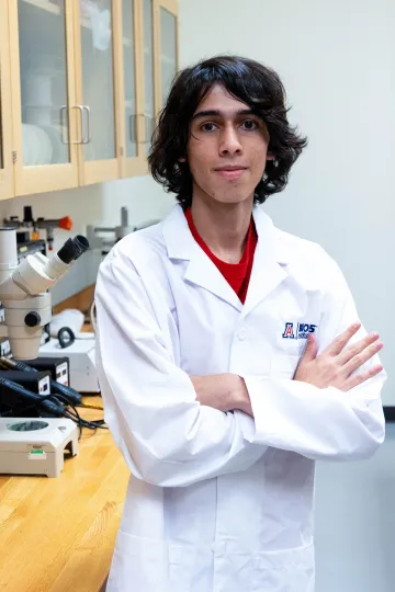 Student in a white lab coat stands confidently with crossed arms in a laboratory setting, smiling at the camera, with a microscope and other scientific equipment in the background.