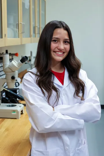 Student in a white lab coat stands confidently with crossed arms in a laboratory setting, smiling at the camera, with a microscope and other scientific equipment in the background.