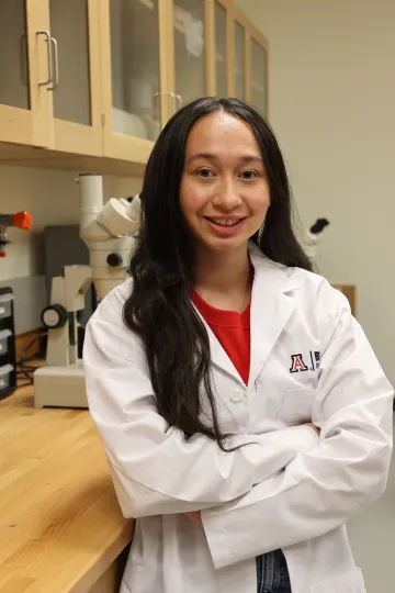 Student in a white lab coat stands confidently with crossed arms in a laboratory setting, smiling at the camera, with a microscope and other scientific equipment in the background.