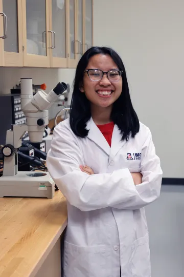 Student in a white lab coat stands confidently with crossed arms in a laboratory setting, smiling at the camera, with a microscope and other scientific equipment in the background.