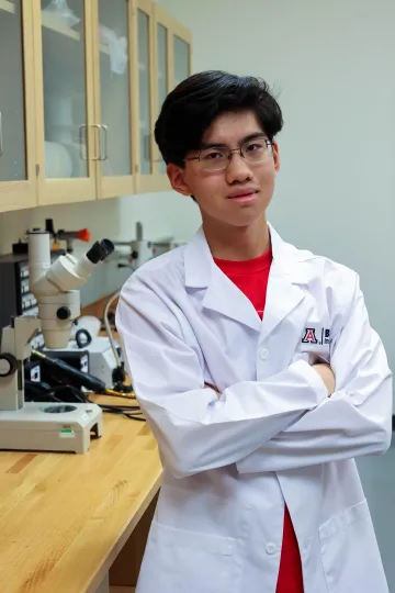 Student in a white lab coat stands confidently with crossed arms in a laboratory setting, smiling at the camera, with a microscope and other scientific equipment in the background.