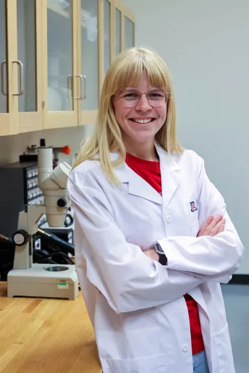 Student in a white lab coat stands confidently with crossed arms in a laboratory setting, smiling at the camera, with a microscope and other scientific equipment in the background.