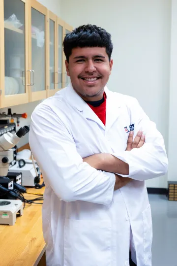 Student in a white lab coat stands confidently with crossed arms in a laboratory setting, smiling at the camera, with a microscope and other scientific equipment in the background.