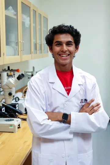 Student in a white lab coat stands confidently with crossed arms in a laboratory setting, smiling at the camera, with a microscope and other scientific equipment in the background.