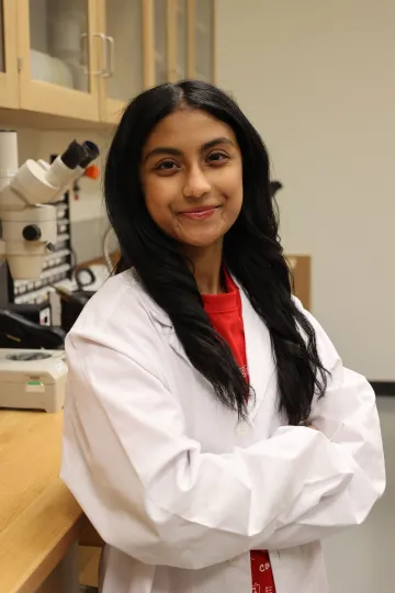 Student in a white lab coat stands confidently with crossed arms in a laboratory setting, smiling at the camera, with a microscope and other scientific equipment in the background.