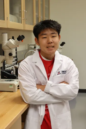 Student in a white lab coat stands confidently with crossed arms in a laboratory setting, smiling at the camera, with a microscope and other scientific equipment in the background.