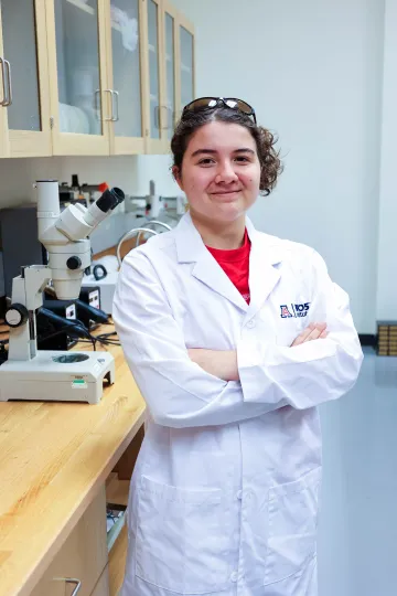 Student in a white lab coat stands confidently with crossed arms in a laboratory setting, smiling at the camera, with a microscope and other scientific equipment in the background.