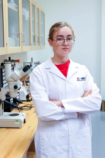 Student in a white lab coat stands confidently with crossed arms in a laboratory setting, smiling at the camera, with a microscope and other scientific equipment in the background.