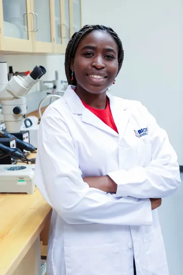 Student in a white lab coat stands confidently with crossed arms in a laboratory setting, smiling at the camera, with a microscope and other scientific equipment in the background.