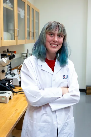 Student in a white lab coat stands confidently with crossed arms in a laboratory setting, smiling at the camera, with a microscope and other scientific equipment in the background.