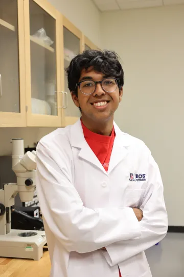 Student in a white lab coat stands confidently with crossed arms in a laboratory setting, smiling at the camera, with a microscope and other scientific equipment in the background.