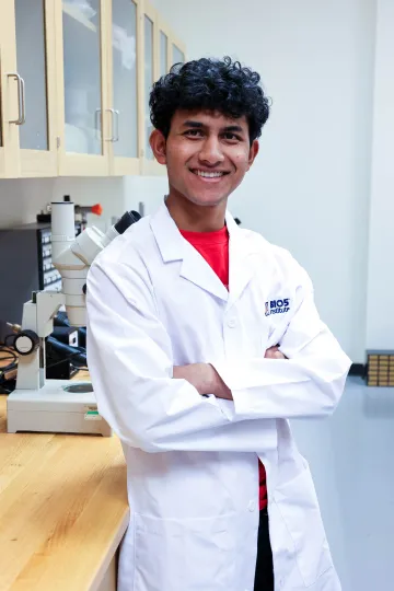 Student in a white lab coat stands confidently with crossed arms in a laboratory setting, smiling at the camera, with a microscope and other scientific equipment in the background.