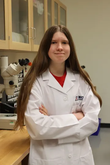 Student in a white lab coat stands confidently with crossed arms in a laboratory setting, smiling at the camera, with a microscope and other scientific equipment in the background.