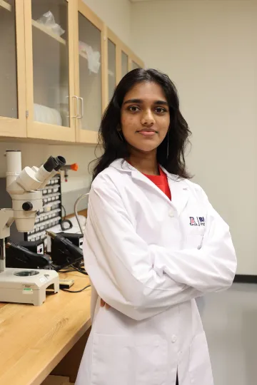 Student in a white lab coat stands confidently with crossed arms in a laboratory setting, smiling at the camera, with a microscope and other scientific equipment in the background.