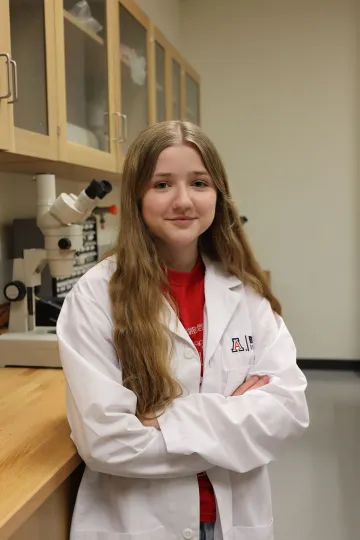 Student in a white lab coat stands confidently with crossed arms in a laboratory setting, smiling at the camera, with a microscope and other scientific equipment in the background.