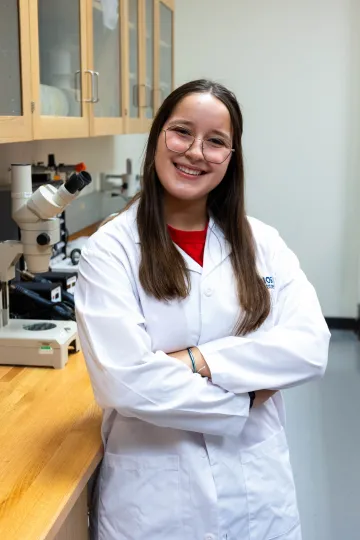 Student in a white lab coat stands confidently with crossed arms in a laboratory setting, smiling at the camera, with a microscope and other scientific equipment in the background.