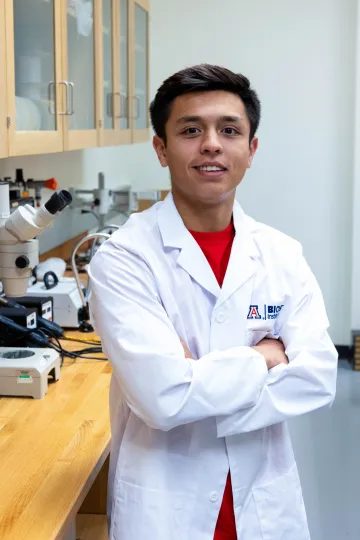 Student in a white lab coat stands confidently with crossed arms in a laboratory setting, smiling at the camera, with a microscope and other scientific equipment in the background.