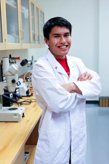 Student in a white lab coat stands confidently with crossed arms in a laboratory setting, smiling at the camera, with a microscope and other scientific equipment in the background.