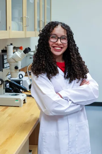 Student in a white lab coat stands confidently with crossed arms in a laboratory setting, smiling at the camera, with a microscope and other scientific equipment in the background.