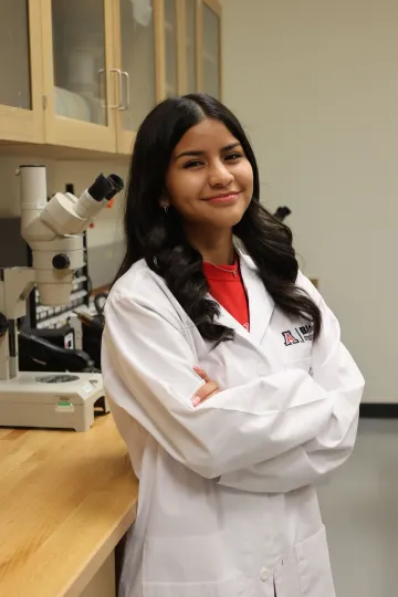 Student in a white lab coat stands confidently with crossed arms in a laboratory setting, smiling at the camera, with a microscope and other scientific equipment in the background.