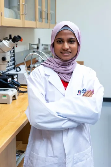 Student in a white lab coat stands confidently with crossed arms in a laboratory setting, smiling at the camera, with a microscope and other scientific equipment in the background.