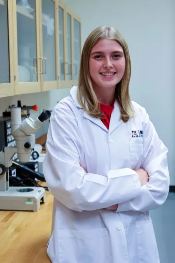 Student in a white lab coat stands confidently with crossed arms in a laboratory setting, smiling at the camera, with a microscope and other scientific equipment in the background.