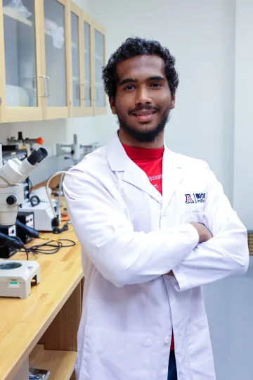 Student in a white lab coat stands confidently with crossed arms in a laboratory setting, smiling at the camera, with a microscope and other scientific equipment in the background.