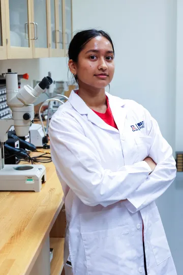 Student in a white lab coat stands confidently with crossed arms in a laboratory setting, smiling at the camera, with a microscope and other scientific equipment in the background.