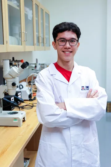 Student in a white lab coat stands confidently with crossed arms in a laboratory setting, smiling at the camera, with a microscope and other scientific equipment in the background.