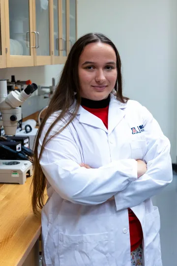 Student in a white lab coat stands confidently with crossed arms in a laboratory setting, smiling at the camera, with a microscope and other scientific equipment in the background.