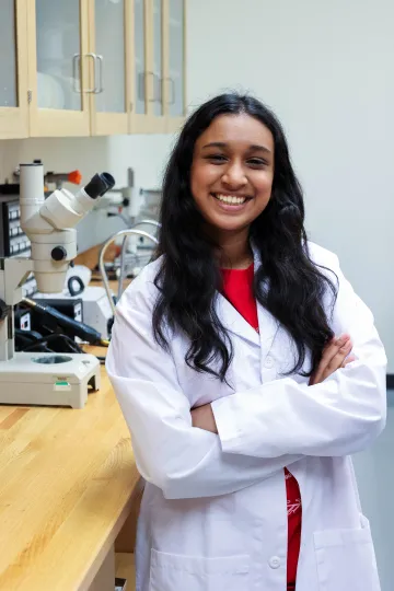 Student in a white lab coat stands confidently with crossed arms in a laboratory setting, smiling at the camera, with a microscope and other scientific equipment in the background.