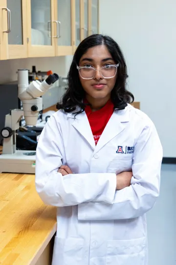 Student in a white lab coat stands confidently with crossed arms in a laboratory setting, smiling at the camera, with a microscope and other scientific equipment in the background.