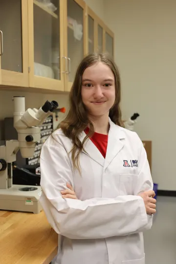 Student in a white lab coat stands confidently with crossed arms in a laboratory setting, smiling at the camera, with a microscope and other scientific equipment in the background.