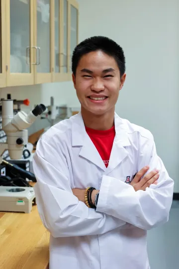 Student in a white lab coat stands confidently with crossed arms in a laboratory setting, smiling at the camera, with a microscope and other scientific equipment in the background.