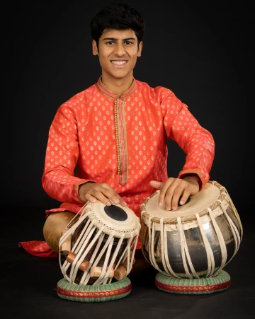 Young person in brightly colored clothing playing Indian hand drums against a black backdrop