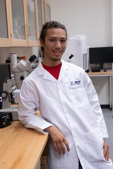 A person in a white lab coat with standing with arms crossed in a laboratory filled with scientific equipment.