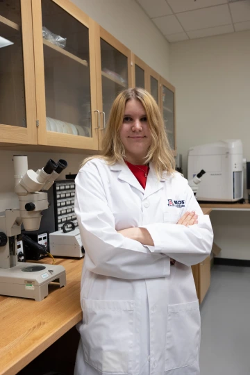 A person in a white lab coat with standing with arms crossed in a laboratory filled with scientific equipment.