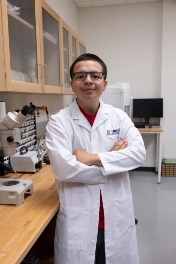 A person in a white lab coat with standing with arms crossed in a laboratory filled with scientific equipment.