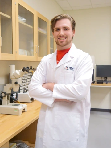 A person in a white lab coat with standing with arms crossed in a laboratory filled with scientific equipment.