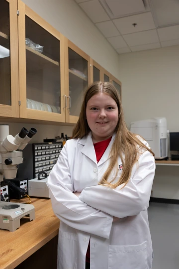 A person in a white lab coat with standing with arms crossed in a laboratory filled with scientific equipment.