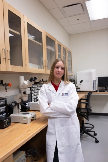A person in a white lab coat with standing with arms crossed in a laboratory filled with scientific equipment.
