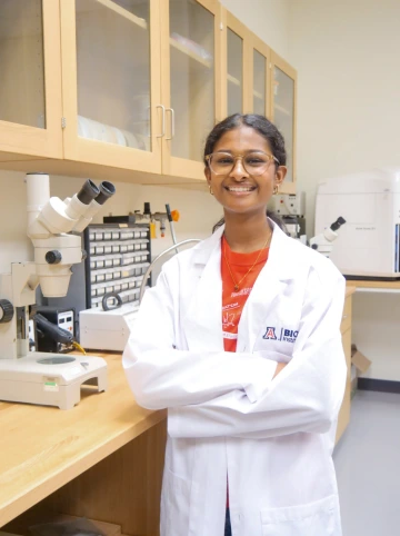 A person in a white lab coat with standing with arms crossed in a laboratory filled with scientific equipment.