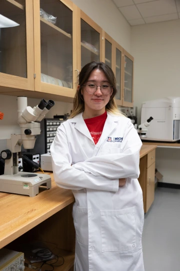 A person in a white lab coat with standing with arms crossed in a laboratory filled with scientific equipment.