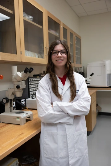 A person in a white lab coat with standing with arms crossed in a laboratory filled with scientific equipment.