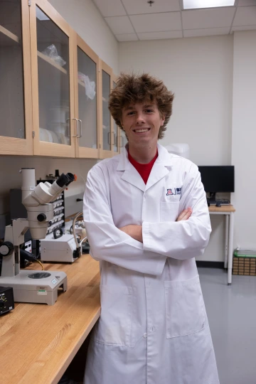 A person in a white lab coat with standing with arms crossed in a laboratory filled with scientific equipment.